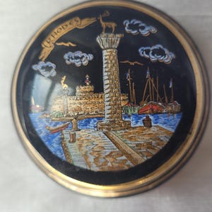 Op de afbeelding: Een ronde, zwarte decoratieve schaal met een gouden rand, met een gedetailleerde illustratie van een kustlandschap. De afbeelding bevat een hoge kolom, boten en het woord "RHODES" in een gebogen banner.