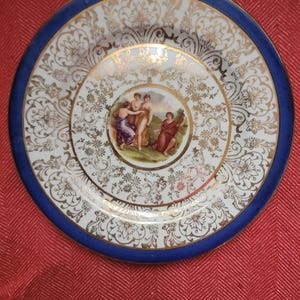 May include: A decorative plate with a blue rim and a white center featuring gold floral patterns. The center of the plate has a painted scene of figures in a garden. A gold border surrounds the central image.