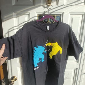 Puede incluir: Camiseta negra con un gráfico de un monstruo azul y un monstruo amarillo luchando. El texto "American Red Cross" está impreso en la parte inferior de la camiseta.