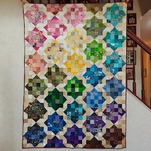 May include: A handmade quilt with a starburst pattern in a rainbow of colors. The quilt features squares of fabric in shades of pink, yellow, green, blue, and purple, set against a neutral background. The quilt is hanging on a wall.