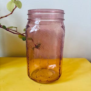 May include: A vintage-style, clear glass jar with a pinkish-purple hue. The jar features embossed floral patterns and a screw-on lid. The jar is sitting on a yellow surface. The jar is a decorative item.