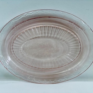 Könnte beinhalten: Eine transparente, ovale Servierplatte mit rosa Farbton. Die Platte hat einen gewellten Rand und zwei Griffe. Zarte Blumenmuster sind am Rand und in der Mitte eingeätzt. Die Platte ist etwa 35,5 cm lang.
