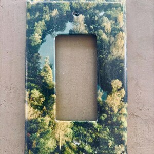 May include: A rectangular light switch cover featuring a scenic forest landscape with green and yellow trees. The cover has a rectangular opening in the center and a small hole at the top. The background is a neutral tan color.
