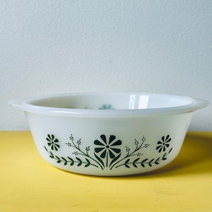 Puede incluir: Una fuente de horno de vidrio opalino blanco con un patrón floral verde oscuro repetido alrededor del exterior. El cuenco tiene una forma redonda con asas laterales integradas y un acabado liso y brillante, mostrando un diseño botánico de flores.