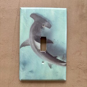 Puede incluir: Una cubierta de interruptor rectangular con un tiburón martillo nadando en un océano azul verdoso. El tiburón es gris y blanco, con una cabeza distintiva en forma de martillo. La cubierta tiene una abertura rectangular.