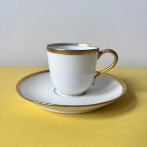 May include: White teacup and saucer set with gold trim. The teacup has a curved handle and a gold band around the rim. The saucer also has a gold rim. The set is on a yellow surface.