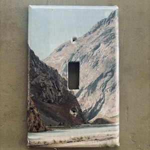 May include: A decorative light switch cover featuring a landscape image of a mountain range and a body of water. The cover has a rectangular shape with a single switch opening.