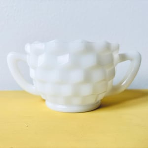 Puede incluir: Un azucarero blanco vintage con un patrón de panal. El cuenco tiene dos asas y un borde ligeramente acampanado. El azucarero está hecho de vidrio opaco y se asienta sobre una superficie amarilla.