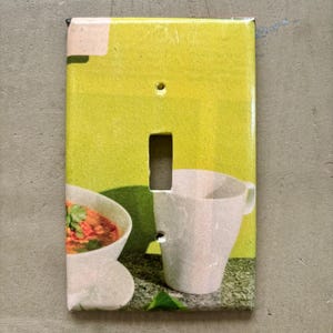 May include: A decorative light switch cover featuring a vibrant design. The cover has a yellow background with a white coffee cup and a bowl of soup. The switch plate has a rectangular opening for the switch and two screw holes.
