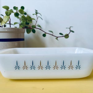 May include: A rectangular, white glass baking dish with a decorative pattern of blue and brown starbursts. The dish is on a yellow surface, with a potted plant in the background.