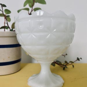 Puede incluir: Un cuenco de postre de cristal blanco de estilo vintage, con borde festoneado y diseño de diamantes. La base del pedestal y el cuenco son de cristal opaco. El cuenco se exhibe sobre una superficie amarilla, con una maceta de cerámica y vegetación en el fondo.