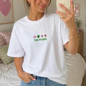 May include: A white short-sleeve t-shirt featuring a small embroidered graphic on the chest. The design includes a pink bow, a green frog, a pink flower, and a red mushroom, with the green text "TEA PLEASE" printed underneath the icons.