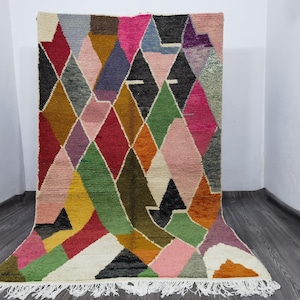 May include: A colorful, geometric rug with a diamond pattern. The rug features a variety of colors, including red, pink, green, yellow, and black. It has a white fringe along the bottom edge. The rug is made of a thick, woven material.
