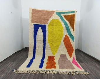 Puede incluir: Una alfombra rectangular de color blanco roto con un diseño geométrico colorido. La alfombra presenta formas en amarillo, azul, naranja, rosa, marrón y turquesa. Tiene un fleco en el borde inferior. La alfombra está hecha de un material grueso y texturizado.