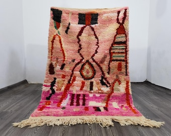 May include: A pink and cream-colored Moroccan rug with a geometric design. The rug features a central diamond-like pattern with red, black, and brown accents. The rug has a fringe along the bottom edge.