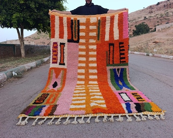 May include: A rectangular wool rug featuring a vibrant, multi-colored geometric pattern with bold blocks of orange, pink, green, blue, and black against a cream background. The rug has a thick, textured pile and a knotted fringe along the bottom edge.