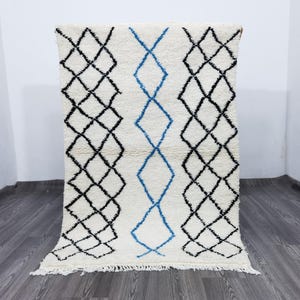 May include: A rectangular, off-white rug with a geometric design. The rug features black diamond patterns on the sides and a central blue diamond pattern. The rug has a fringe along the bottom edge.