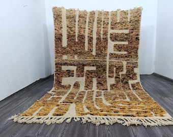 Puede incluir: Una alfombra rectangular con un patrón geométrico en tonos marrón y crema. La alfombra tiene flecos en el borde inferior. El diseño presenta una serie de líneas verticales y horizontales que crean una apariencia texturizada.