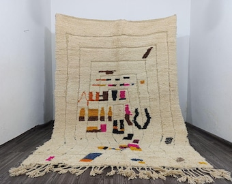 Puede incluir: Alfombra rectangular de color crema con un diseño geométrico. La alfombra presenta un patrón central de cuadrados y rectángulos en tonos marrón, negro, naranja, rosa y azul. La alfombra tiene flecos en el borde inferior.