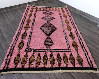Puede incluir: Una alfombra rectangular rosa con un diseño central de diamantes negros. La alfombra tiene un borde negro y está decorada con formas repetidas de diamantes y triángulos negros y dorados. La alfombra mide aproximadamente 152 cm de largo y 91 cm de ancho.