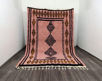 May include: A pink Moroccan rug with a geometric design. The rug features a central diamond pattern in black, surrounded by black and gold diamond shapes. The rug has a fringe along the bottom edge.