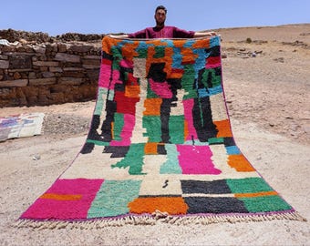 Puede incluir: Una alfombra grande y colorida con un diseño geométrico. La alfombra presenta un mosaico de colores brillantes que incluyen rosa, verde, naranja y negro. La alfombra se exhibe al aire libre, mostrando su tamaño y textura.