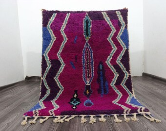 Puede incluir: Una alfombra rectangular tejida a mano con un diseño vibrante. La alfombra presenta un campo magenta intenso con patrones geométricos en azul, morado y verde azulado. Los bordes están adornados con borlas. La alfombra mide aproximadamente 152 cm por 183 cm.