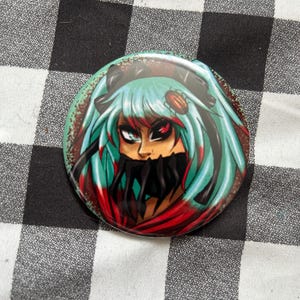 May include: A circular pin-back button featuring a stylized illustration of a character with teal hair, a black mask covering the mouth, and one red eye. The character has a small brown insect on their hair, set against a dark red background.