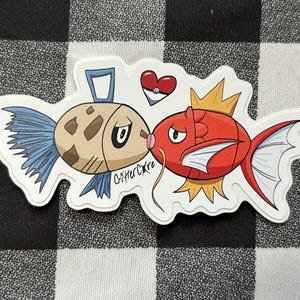Puede incluir: Una pegatina troquelada que muestra un pez redondo de color tostado con aletas azules y un pez rojo de aletas puntiagudas besándose. Arriba hay un icono de Poké Ball en forma de corazón. El texto "CritterCore" está escrito en negro entre ambos.