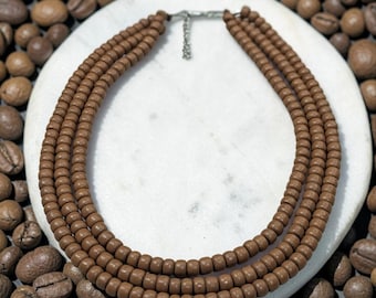 May include: A multi-strand necklace featuring three rows of small, round, matte brown beads. The necklace is secured with a silver-toned metal clasp and a short adjustable chain link at the back, creating a layered appearance with uniform bead sizing.