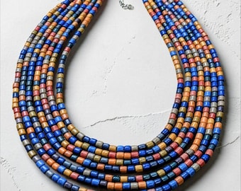 May include: A multi-strand necklace featuring six rows of cylindrical beads in alternating shades of blue, orange, yellow, and grey. The strands are connected by a silver-toned metal clasp with an adjustable chain link extension at the top centre.