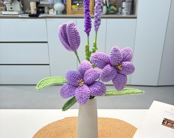 May include: A handmade bouquet of crochet flowers in a white vase. The arrangement includes purple tulips, lavender, and other purple flowers with green leaves. The flowers are displayed on a round, woven table mat.