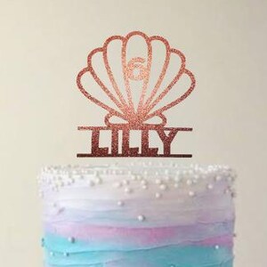 Op de afbeelding: Een verjaardagstaart met blauwe, roze en witte gradiënt frosting, bekroond met een glitterende roségouden zeeschelp taarttopper met de tekst "LILLY" en het nummer "6". De taart staat op een witte taartstandaard.