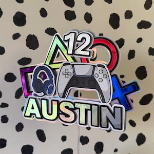 May include: A custom cake topper featuring the name AUSTIN and the number 12 in a holographic finish. The design includes a white and black game controller, blue gaming headphones, and the iconic triangle, circle, square, and cross gaming symbols.