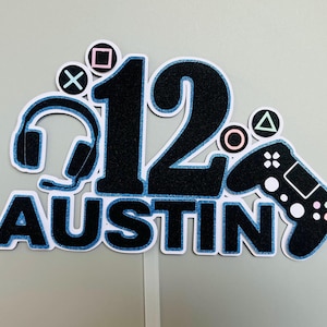 May include: A black and blue cake topper with the number 12, the name Austin, and gaming symbols including a headset, a controller, and a triangle, square, and X.