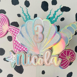 May include: A mermaid-themed cake topper featuring a pink mermaid tail, a seashell, and the name "Nicola". The number "3" is displayed on the seashell. The topper has iridescent and glitter accents, ideal for a birthday celebration.