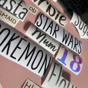 Puede incluir: Una colección de pegatinas de vinilo blanco con letras negras e iridiscentes. Las pegatinas presentan varias palabras y frases, incluyendo "Pokemon", "Star Wars", "Mum" y "Flour". El número "18" también es visible.