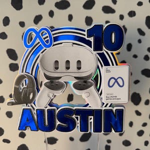 May include: A layered cake topper featuring a white virtual reality headset, two controllers, a black gaming headset, and a Meta Quest gift card. The design includes the number 10, the name AUSTIN in blue metallic letters, and the Meta infinity logo.