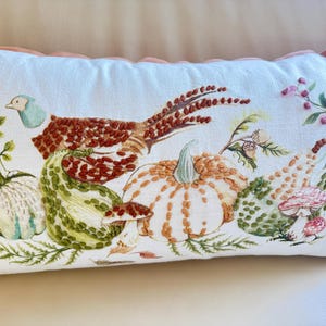 Pu&ograve; includere: Cuscino rettangolare bianco con ricamo a tema raccolto autunnale. Presenta un fagiano, zucche, zucche e funghi nei toni del marrone, verde, arancione e rosa. Il cuscino ha un bordo smerlato.