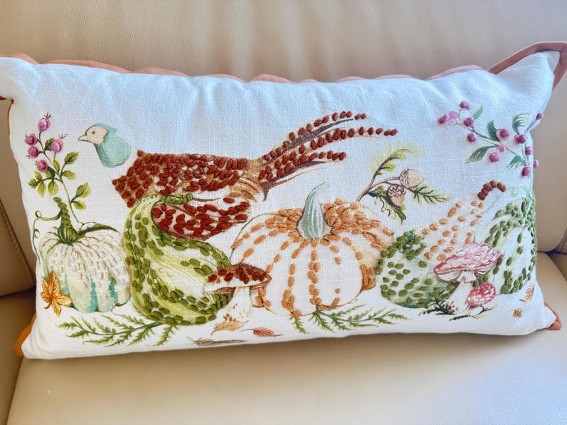 Pu&ograve; includere: Cuscino rettangolare bianco con ricamo a tema raccolto autunnale. Presenta un fagiano, zucche, zucche e funghi nei toni del marrone, verde, arancione e rosa. Il cuscino ha un bordo smerlato.