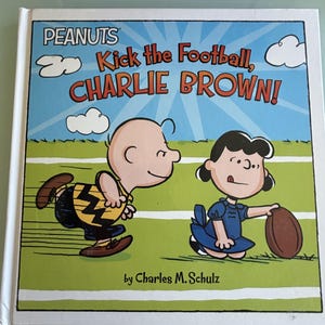 May include: A book cover featuring Charlie Brown and Lucy from the Peanuts comic strip. The title reads "Kick the Football, Charlie Brown!" with a blue sky background and green field. The author's name, Charles M. Schulz, is at the bottom.