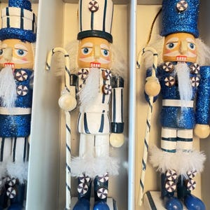 May include: Three decorative wooden nutcrackers in a box. The nutcrackers have blue and white uniforms with glitter accents. Each figure holds a candy cane-striped staff with a white ball at the end. The nutcrackers have white beards and hats.