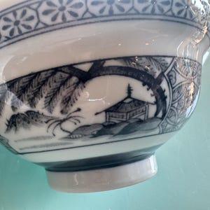 May include: White ceramic bowl with a blue and white landscape design. The bowl features a detailed illustration of a building and trees, with a floral pattern around the rim. The bowl has a rounded shape and a small foot.