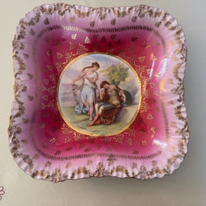 May include: Square, antique decorative dish with a ruffled edge. The dish features a vibrant pink and gold color scheme. The center of the dish has a circular painting of two figures in classical attire. The dish is likely made of porcelain.