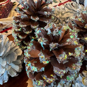 May include: Three pine cones decorated with iridescent glitter. The pine cones are brown and have a natural texture. The glitter is a mix of blue, green, and silver.