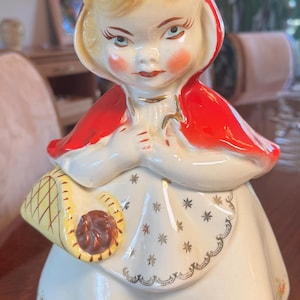 May include: A ceramic cookie jar shaped like a young person wearing a bright red hood and cape. The figure has blonde hair, rosy cheeks, and holds a yellow basket with a brown item inside. The white dress features gold star accents and a floral hem.