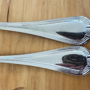 May include: Two polished silver spoons with a decorative, ridged design on the handle ends. The spoons are set against a light brown wooden surface, reflecting the surrounding environment.