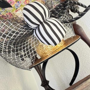 May include: A decorative headband featuring a miniature gold glitter top hat with a black brim. The hat is adorned with a large bow with black and white stripes. Black netting and floral fabric add to the whimsical design.
