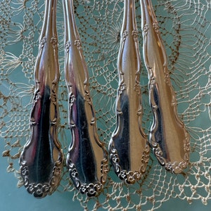 May include: Four silver spoons with ornate floral designs on the handles. The spoons are arranged in a row on a white crocheted doily.