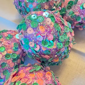 May include: Close-up of several round, decorative ornaments. Each ornament is covered in a variety of colorful sequins, including pink, green, and orange. The base color of the ornaments is a light purple.